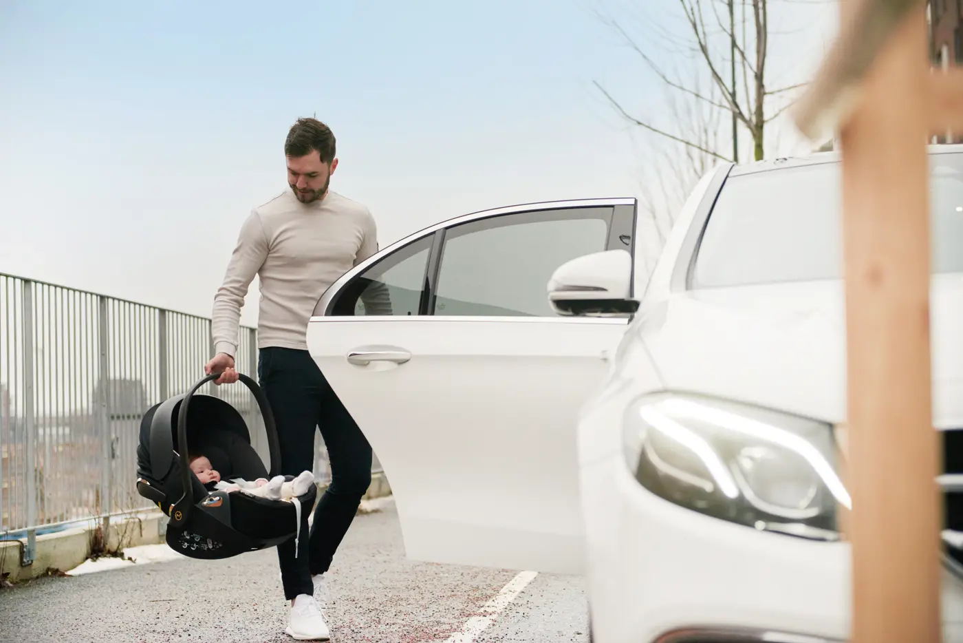 En man bär ett babyskydd med en bebis i, intill en vit bil.